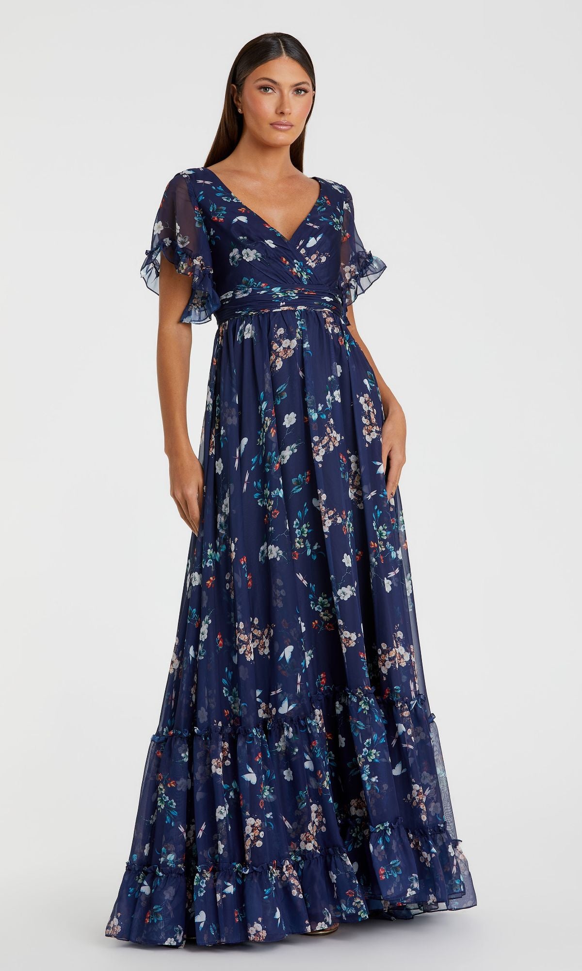 A woman models the Mac Duggal 70241 Floral-Print Long Formal Dress in navy blue with short ruffled sleeves, V-neckline, and tiered hem, standing against a plain light background.
